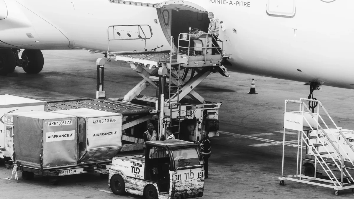 A white airplane with a cargo loading ramp and a truck with a lift gate, positioned in front of the plane, with the ramp extended and the tr