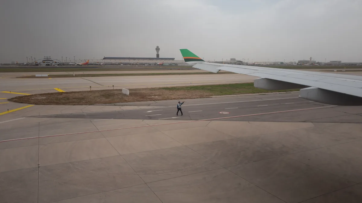 An airport runway with a plane's wing visible on the right side, a person standing in the middle of the runway, and a control tower in the background.
