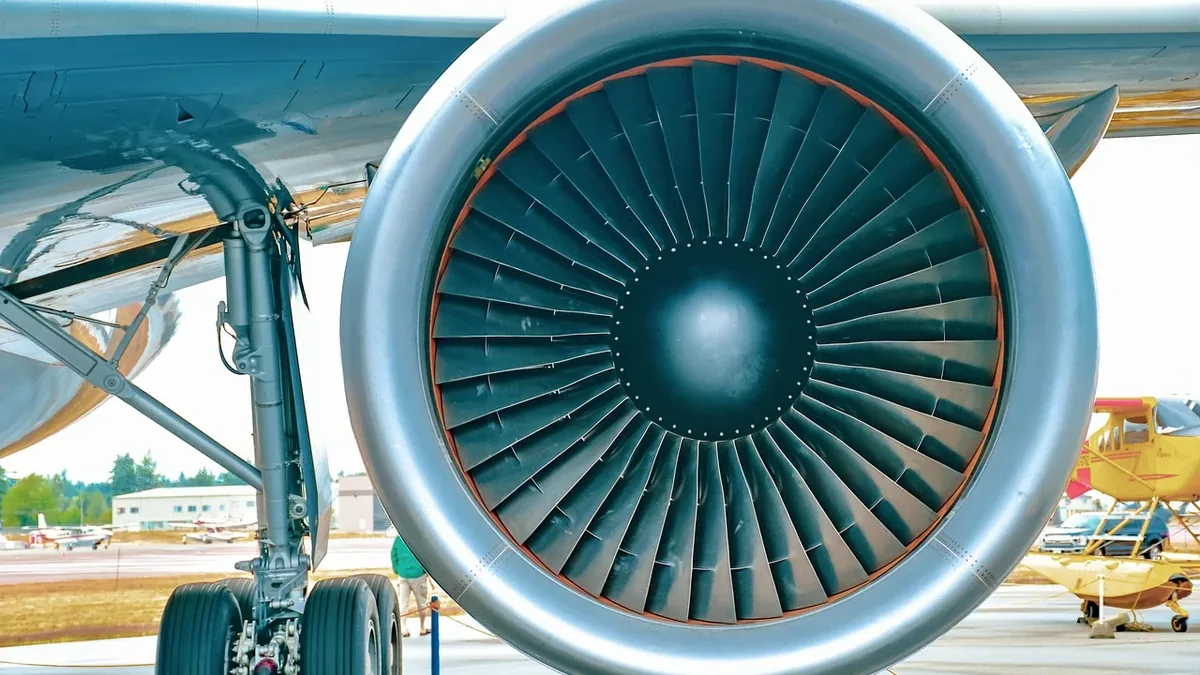 A large airplane engine with a fan and blades, attached to the wing of an airplane on the ground.