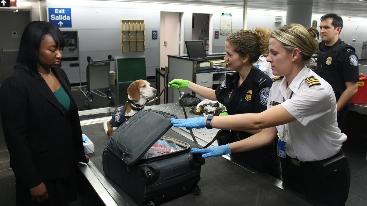 A woman is being screened by two TSA agents and a dog at an airport security checkpoint.